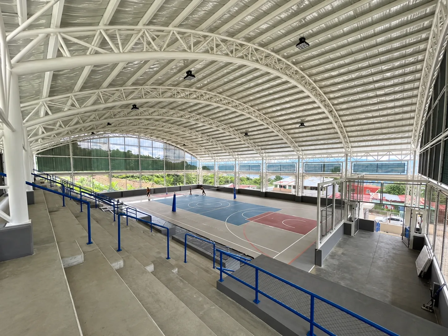 Gimnasio techado con estructura metálica curva, cancha multiuso y graderías de concreto
